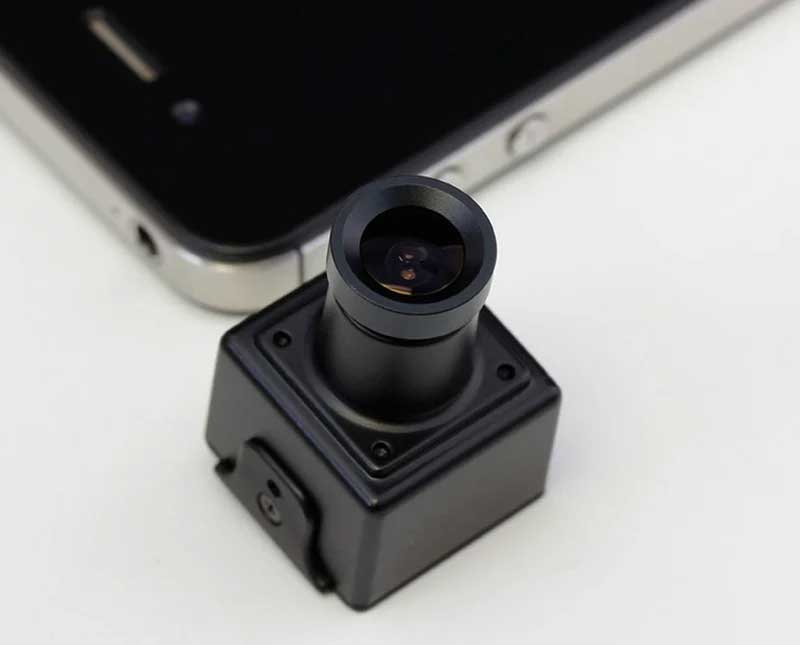 A small black square camera module with a cylindrical lens assembly, shown in close-up on a white surface with part of a smartphone visible in the background.
