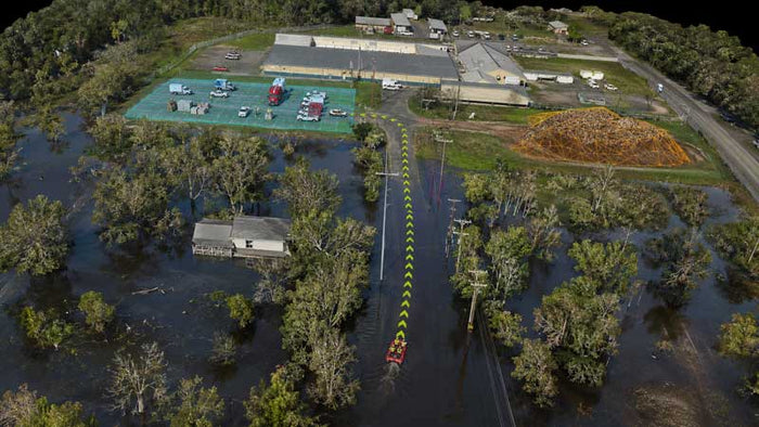 3D Photogrammetry view of a flooded area with trees, a small building, a parking lot with several vehicles, industrial buildings, a pile of wood, and a narrow road with a vehicle driving through deep water.