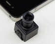A small black square camera module with a cylindrical lens assembly, shown in close-up on a white surface with part of a smartphone visible in the background.
