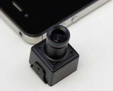 A small black square camera module with a cylindrical lens assembly, shown in close-up on a white surface with part of a smartphone visible in the background.