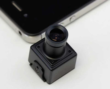 A small black square camera module with a cylindrical lens assembly, shown in close-up on a white surface with part of a smartphone visible in the background.