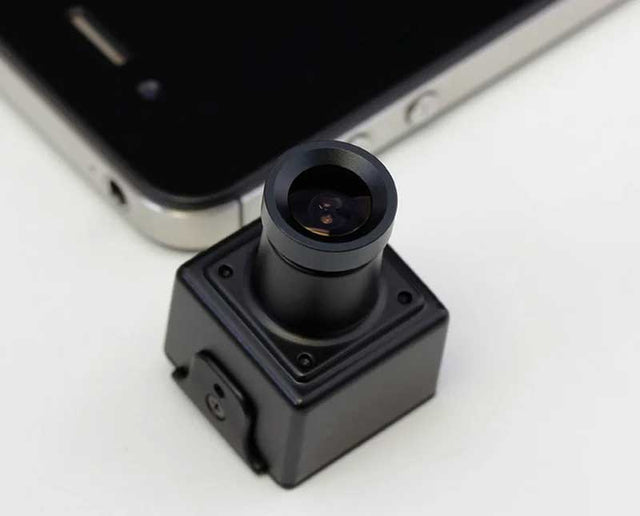 A small black square camera module with a cylindrical lens assembly, shown in close-up on a white surface with part of a smartphone visible in the background.