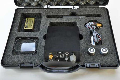 An covert body wire kit displayed inside its custom foam-lined black carrying case. The kit includes a central black device with control knobs, a gold-colored battery, a small rectangular component, a coiled cable with connectors, and two circular metal parts.