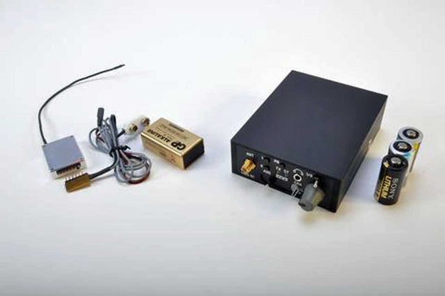 A black police body wire with attached wires and a small gold-colored box, shown alongside two batteries on a plain white background.