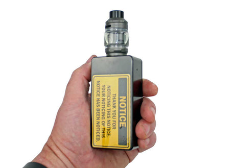 A hand holding a covert video vape device with a yellow and black 'Notice' label, shown against a plain white background.