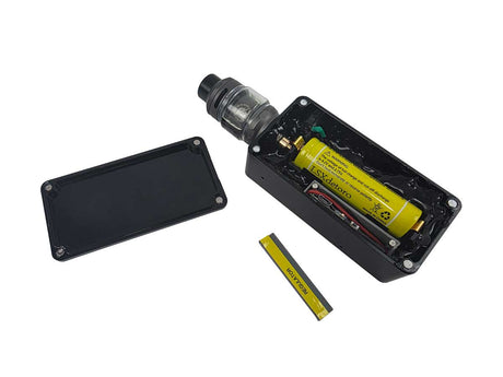 A black undercover video camera device with its lid removed, showing a yellow cylindrical battery inside, alongside a separate black lid and a small yellow accessory on a white background