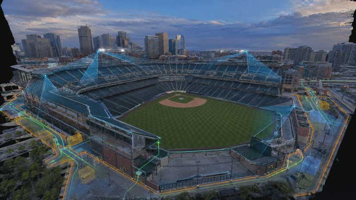 Drone photogrammetry view of a baseball stadium at dusk with a city skyline in the background, showing the field, stands, lighting, and surrounding urban environment.