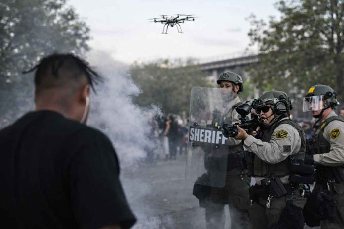 Law enforcement officers in riot gear with a drone flying above during a protest.